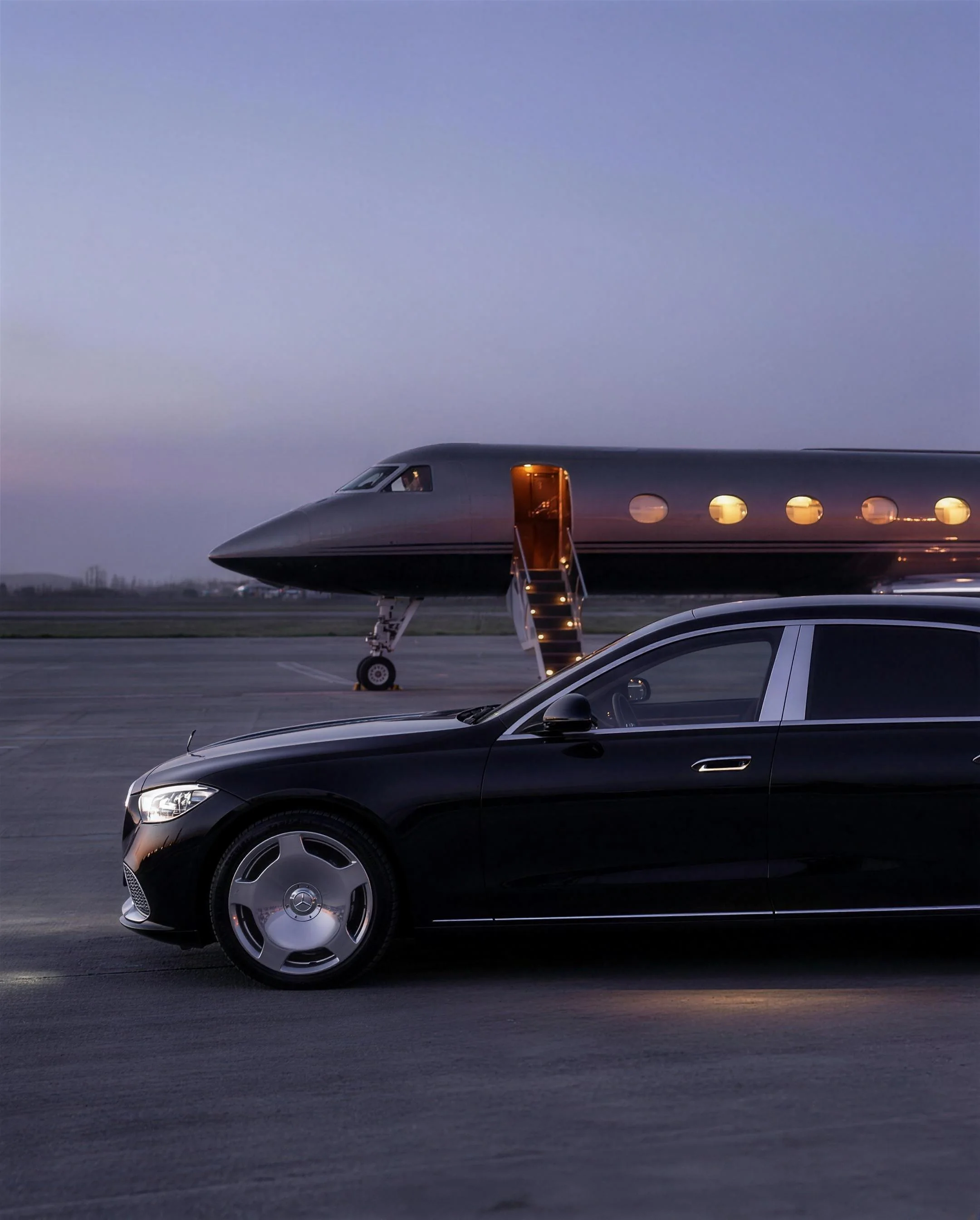 A chauffeured Maybach sedan waiting on the tarmac beside a lit private jet at dusk