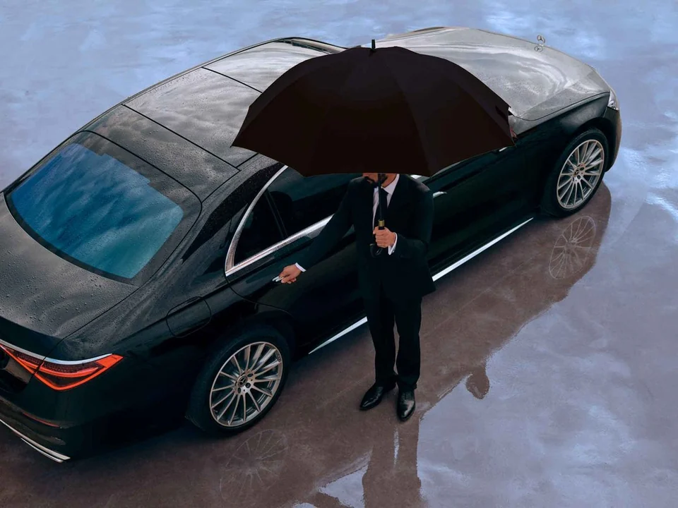 A chauffeur holding an umbrella beside a black sedan in the rain, reaching for the rear door